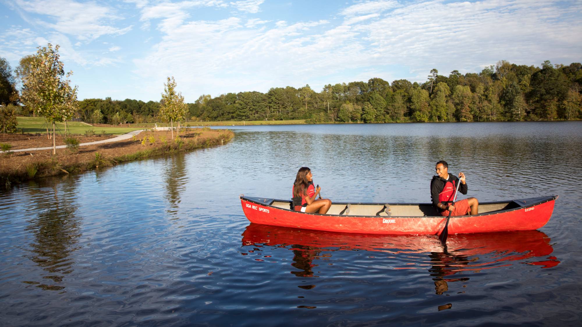 Two people smile at each other while paddling a canoe on a lake under a partly cloudy blue sky