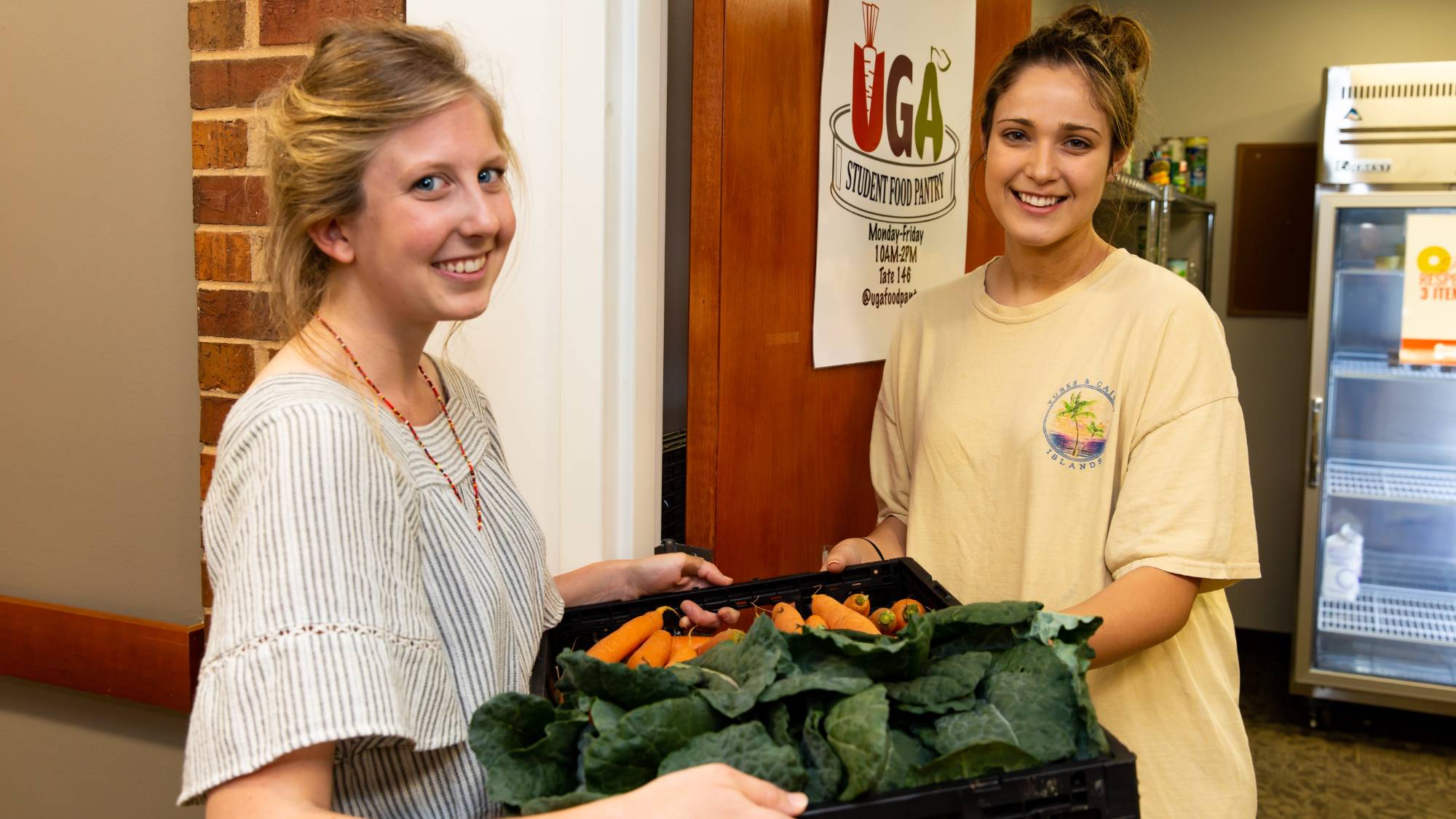 Two young women smile while one hands a tray of fresh vegetables to the other in a doorway labeled UGA Food Pantry